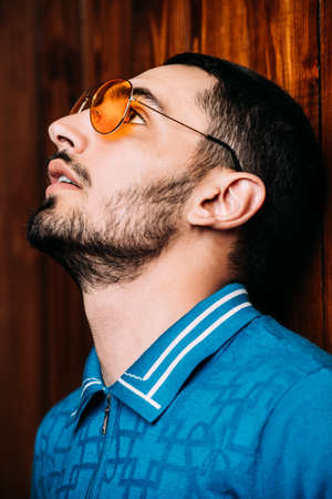 Vogue shot of a handsome male model in a T-shirt and yellow sunglasses standing by a wooden wall. Men's beauty, fashion. の写真素材