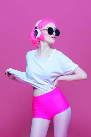 Trendy girl with pink hair wearing sunglasses enjoys the music on headphones. Pink background. Youth style, leisure.の写真素材
