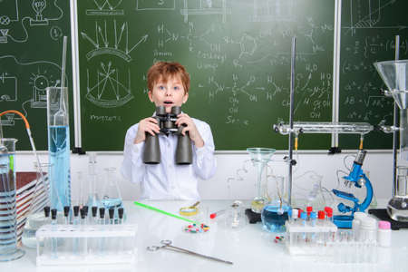 Schoolboy looks through binoculars by a chalkboard on a chemistry lesson. Educational concept.の写真素材