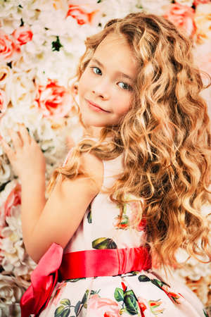 Portrait of a cute little girl in a beautiful summer dress with blonde curly hair. Background of roses. Kid's fashion, style.の写真素材