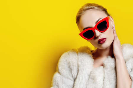 Gorgeous blonde woman posing in luxurious fur coat and pin-up sunglasses. Yellow background. Fashion, beauty. Studio shot.の写真素材
