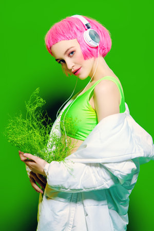 Trendy vivid girl with pink hair wearing headphones holding a bunch of grass. Green background. Beauty, fashion, youth style.の写真素材