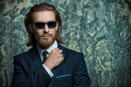 Fashion shot. Handsome young man posing in elegant suit and sunglasses over dark background. Men's beauty, fashion.の写真素材