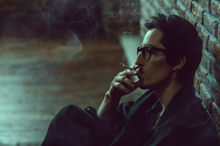 Attractive young man thoughtfully and calmly smoking a cigarette. Loft style interior. Men's beauty, fashion. Optics style.の写真素材