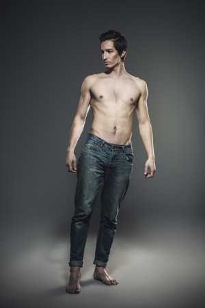 Portrait of a sexy shirtless young man with slender body posing at studio in jeans by the brick wall.  Men's health.の写真素材