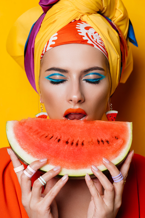 Fashionable woman with bright make-up eating watermelon. Yellow background. Beauty, fashion, make-up concept.の写真素材