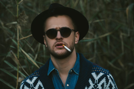 Close-up portrait of a handsome brutal man in cardigan and a hat smoking a cigarette outdoor over autumn nature background. Autumn fashion. Male fashion.の写真素材
