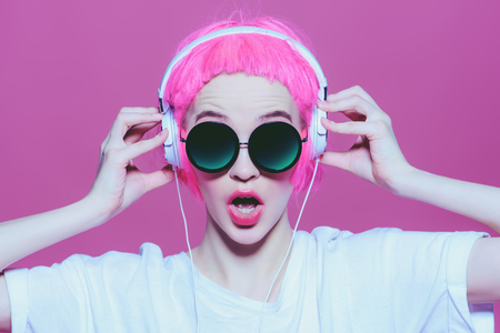 Trendy girl with pink hair wearing sunglasses enjoys the music on headphones. Pink background. Youth style, leisure.の写真素材