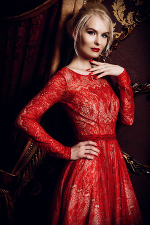 Attractive young woman in evening red dress stands in a luxury apartment. Classic vintage interior. Beauty, fashion. Evening makeup and hairstyle.の写真素材