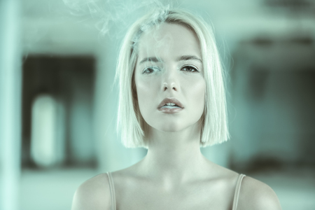 Attractive young girl smokes a cigarette against the backdrop of an abandoned house.の写真素材