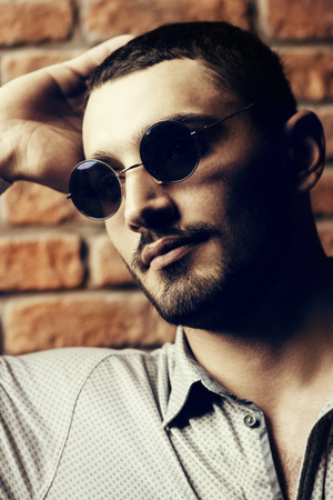 Close-up portrait of a handsome brunet man in sunglasses over brick wall background. Men's beauty, fashion. Men's barbershop, Hairstyle.の写真素材