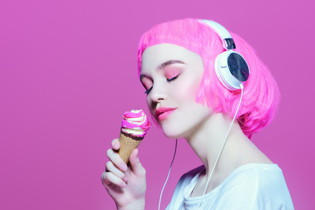 Trendy girl with pink hair wearing headphones is eating ice cream. Pink background. Youth style, leisure.の写真素材