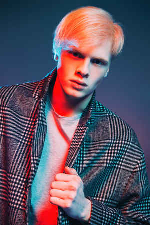 Portrait of a handsome young man with blond hair wearing in a plaid coat, posing at studio over gray background. Men's beauty and health.の写真素材
