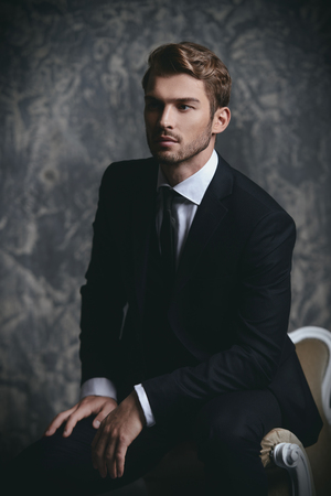 Portrait of a handsome man in an elegant suit sitting in an armchair on a grunge background. Studio shot.の写真素材
