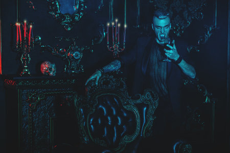 A vampire man is standing in a dark room and drinking wine or blood. Halloween. Beauty, fashion, horror.の写真素材