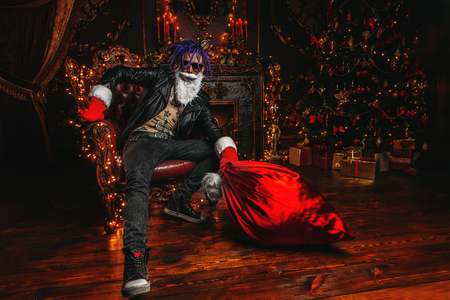 Ð¡heerful punk Santa with a bag of gifts in his hands  in luxurious apartments decorated for Christmas. Bad Santa concept.の写真素材