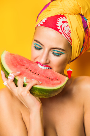 Fashionable woman with bright make-up eating watermelon. Yellow background. Beauty, fashion, make-up concept.の写真素材