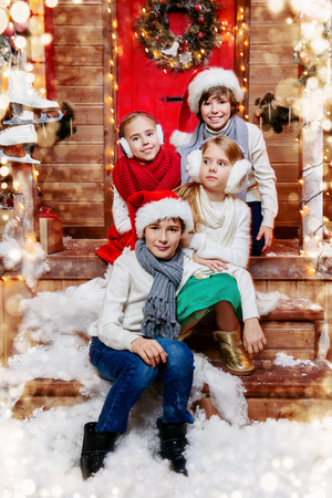 Four cheerful children in winter clothes sit on the porch of the house decorated for Christmas. Kid's fashion. Time for miracles. Merry Christmas and Happy New Year.の写真素材