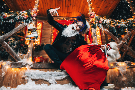 Ð¡heerful punk Santa fools around near his house with a bag of gifts in his hands.の写真素材
