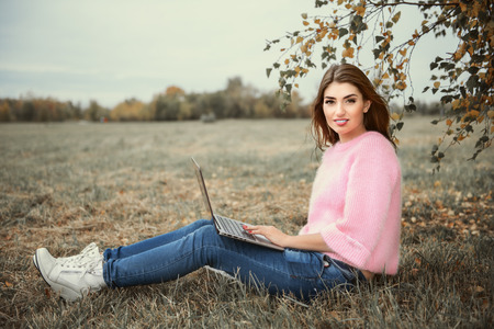 A beautiful young woman is sitting on the grass in the coutryside with a laptop. Lifestyle, autumn fashion. Nature and gadgets.の写真素材