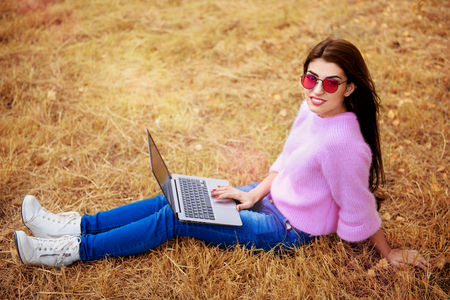 A beautiful young woman is sitting on the grass in the coutryside with a laptop. Lifestyle, autumn fashion. Nature and gadgets.の写真素材