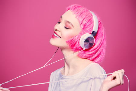Trendy girl with pink hair enjoys the music on headphones. Pink background. Youth style, leisure.の写真素材