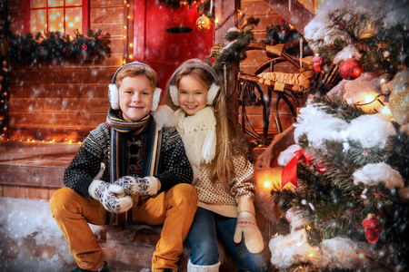 A portrait of children sitting on the porch of their house together. Merry Christmas, happy New Year. Yard with a christmas tree, lights and decorations. Miracle time.の写真素材