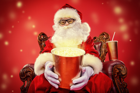 Merry Christmas and Happy New Year. Santa Claus sitting on his armchair and eating popcorn and drinking soda. Entertainment and cinema concept. Red background.の写真素材
