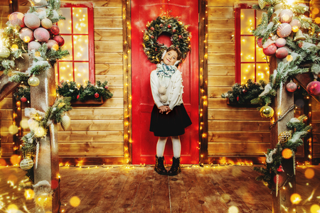 A cute girl is standing on the porch of the house decorated for Christmas and dreaming. Merry Christmas, Happy New Year. Miracle time.の写真素材
