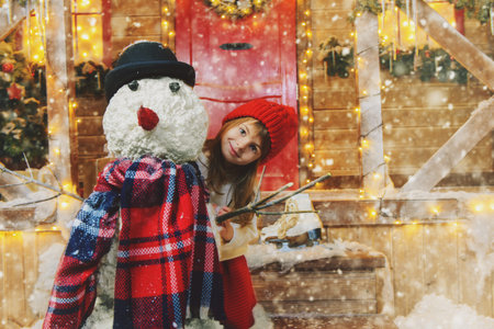 Cute child girl sculpts a snowman near her house decorated for Christmas. Merry Christmas and Happy New Year.の写真素材