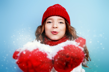 A portrait of a pretty young girl wearing a hat, a sweater, a scarf and mittens, blowing on the snow in her palms. Winter fashion for kids, beauty.の写真素材