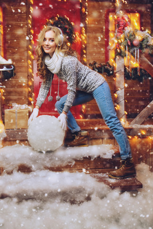 Pretty girl in winter clothes made a big snowball for a snowman near house decorated for Christmas. Merry Christmas and Happy New Year.の写真素材