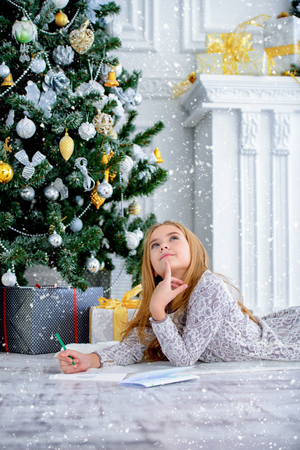 Cute girl  is lieing and writing a letter to Santa Claus near the Christmas tree. Christmas concept. Believe in miracles.の写真素材