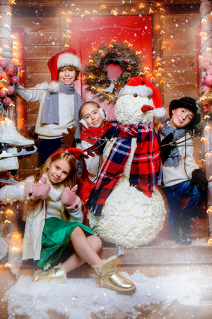 Cheerful children in winter clothes and accessories stand near the snowman on the porch of the house decorated for Christmas. Time for miracles. Merry Christmas and Happy New Year.の写真素材