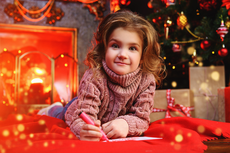 A happy girl is lying in the floor in the room decorated for Christmas and writing a wish letter to Santa. Christmas, New Year.の写真素材