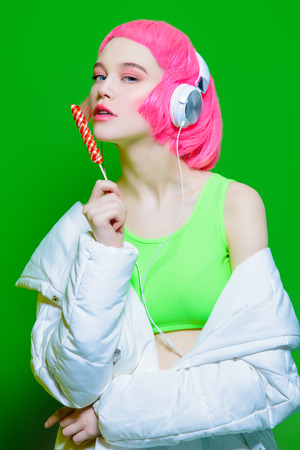Trendy girl with pink hair wearing headphones is eating lollipop. Green background. Youth style, leisure.の写真素材