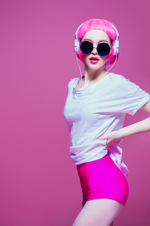 Trendy girl with pink hair wearing sunglasses enjoys the music on headphones. Pink background. Youth style, leisure.の写真素材