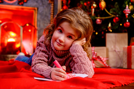 A happy girl is lying in the floor in the room decorated for Christmas and writing a wish letter to Santa. Christmas, New Year.の写真素材
