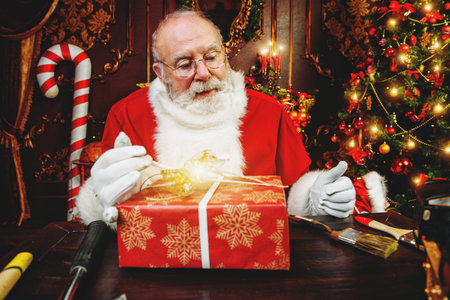 Santa Claus is sitting at a table at home with a gift box. Merry Christmas and Happy New Year. Miracle time.の写真素材
