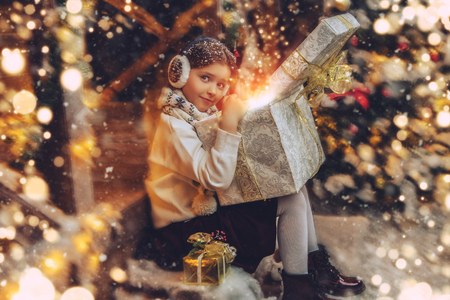 A cute girl is sitting on the porch of the house decorated for Christmas and holding a gift box. Merry Christmas, Happy New Year. Miracle time.の写真素材