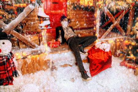 Drunk punk Santa is lying with a bottle and a bag of gifts on the porch of the house, decorated for Christmas.の写真素材