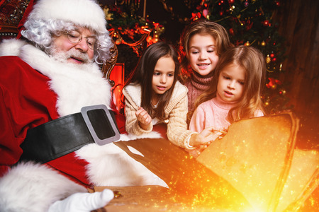 Happy young girls are sitting with Santa Claus at home. Merry Christmas and Happy New Year. Miracle time.の写真素材