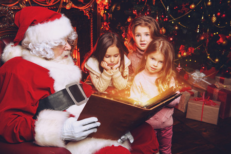 Happy young girls are sitting with Santa Claus at home. Merry Christmas and Happy New Year. Miracle time.の写真素材