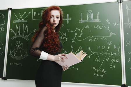Pretty student girl with long red hair posing in school clothes by the chalkboard.の写真素材