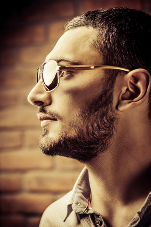 Close-up portrait of a handsome brunet man in sunglasses over brick wall background. Men's beauty, fashion. Men's barbershop, Hairstyle.の写真素材