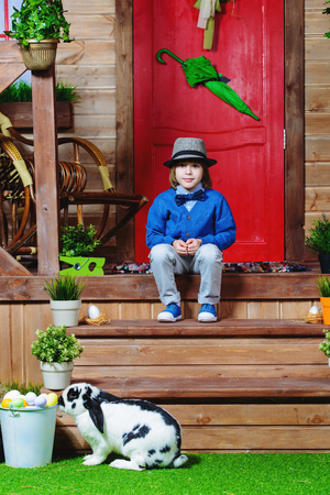 Happy little boy with bunny sitting on the porch near the wooden house. Easter holiday. Rural style, easter decoration.の写真素材