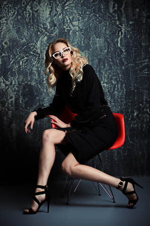 Elegant blonde young woman wearing black dress is sitting on a red chair. Beauty, fashion concept. Full length portrait.の写真素材