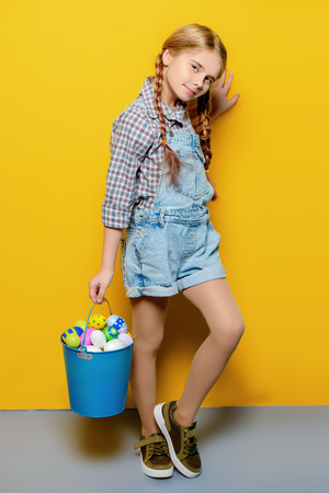 Children's fashion. Cute nine year old girl with long blonde hair is posing in summer clothes with a bucket of easter eggs. Studio shot. Full length portrait.の写真素材