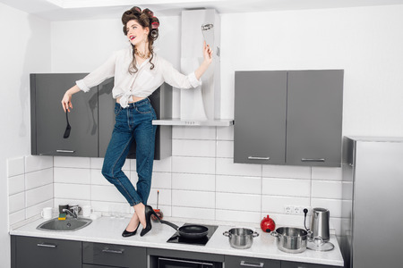 An attractive girl in everyday clothes and curlers stands on the countertop of the kitchen set holding kitchen utensils. Fashion home shot. Full length portrait.の写真素材