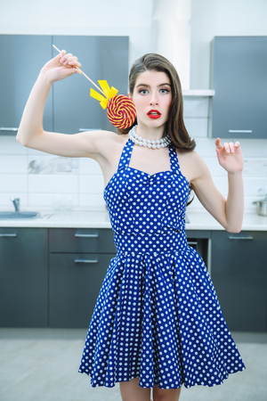 Portrait of attractive young woman in a blue dress with white dots holding a bright Lollipop in his hand on kitchen background. Pin-up style.の写真素材